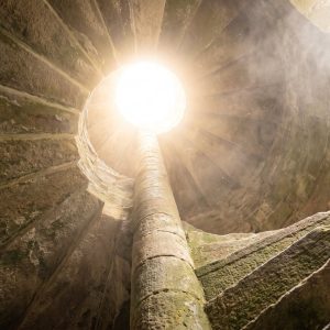 Un escalier de pierre en colimaçon monte vers une ouverture baignée de lumière — abonnement Sādhana