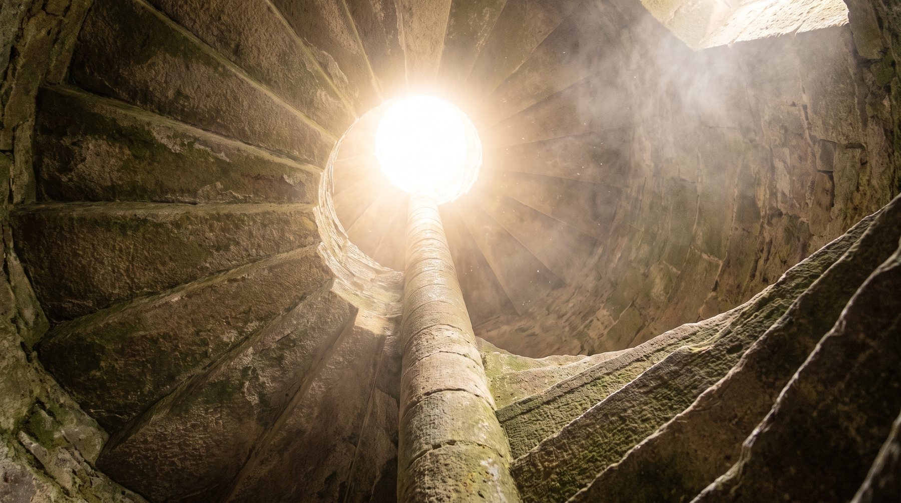 Un escalier de pierre en colimaçon monte vers une ouverture baignée de lumière — abonnement Sādhana