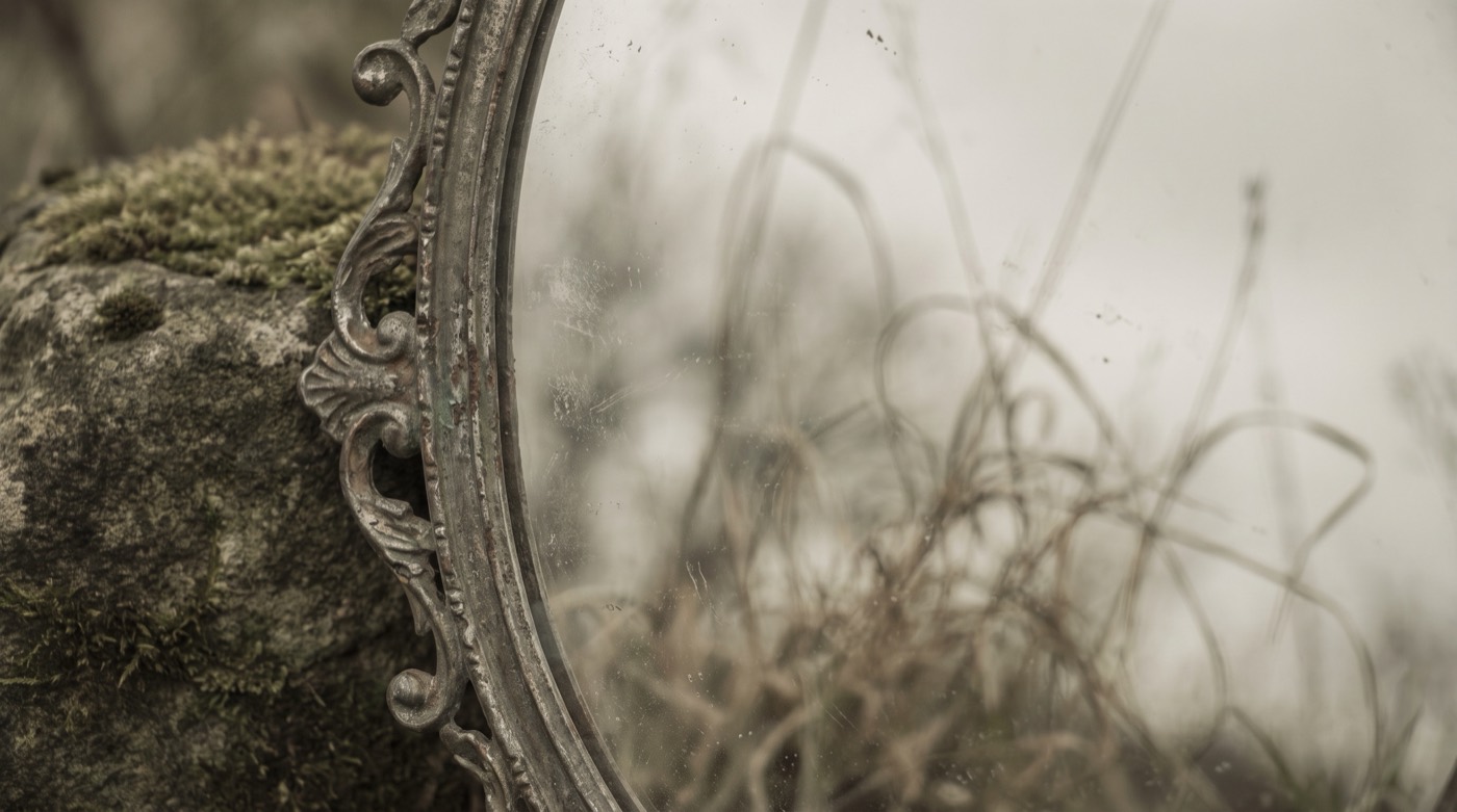 Qui suis-je : un miroir ancien posé contre une pierre, qui reflète un ciel voilé et des herbes sèches